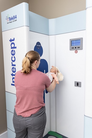 A woman stands outside the scanner, holding a baby horizontally in the scanner, supporting its head with her hands, and body with her arms. 